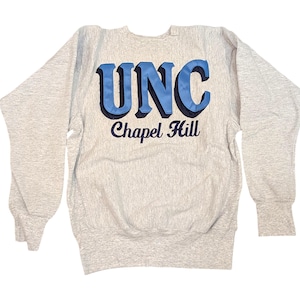 K&ouml;nnte beinhalten: Ein hellgraues Sweatshirt mit dem Text "UNC Chapel Hill" in blauen und schwarzen Buchstaben. Das Sweatshirt hat lange &Auml;rmel und gerippte B&uuml;ndchen und Saum.