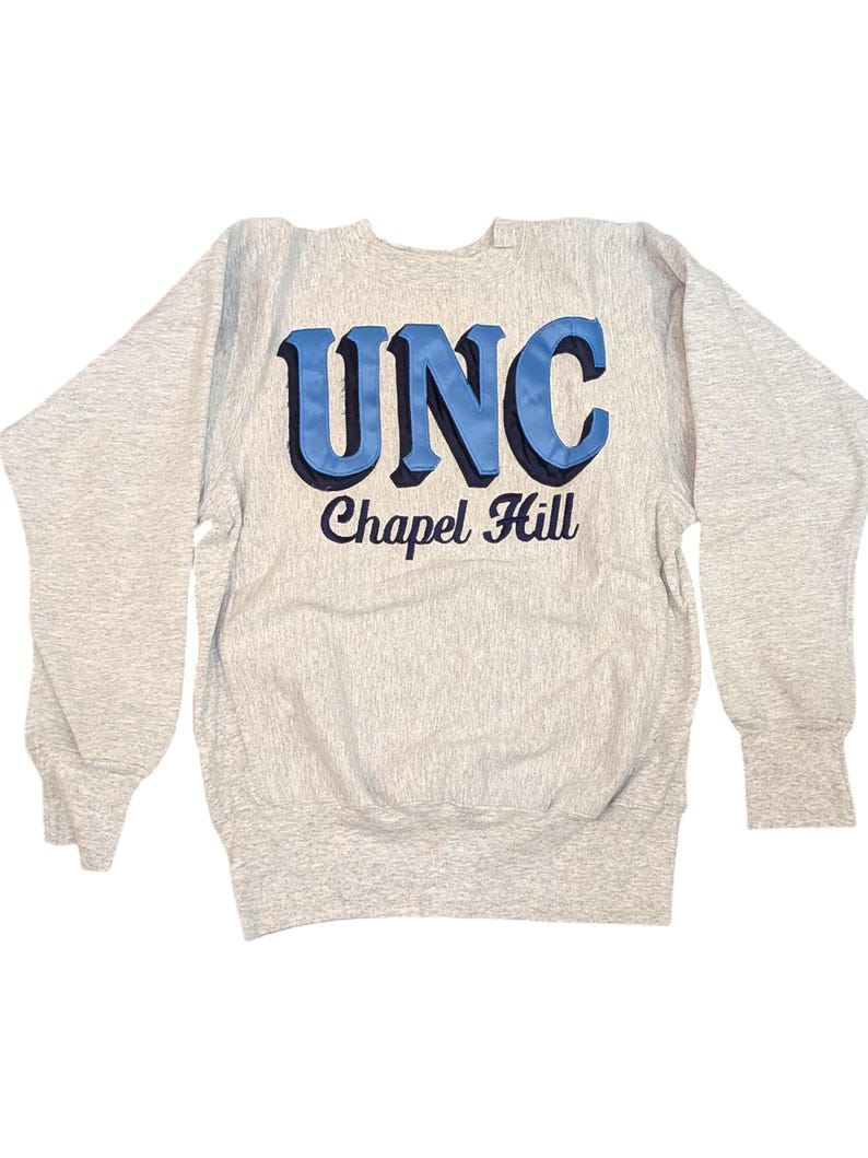 K&ouml;nnte beinhalten: Ein hellgraues Sweatshirt mit dem Text "UNC Chapel Hill" in blauen und schwarzen Buchstaben. Das Sweatshirt hat lange &Auml;rmel und gerippte B&uuml;ndchen und Saum.