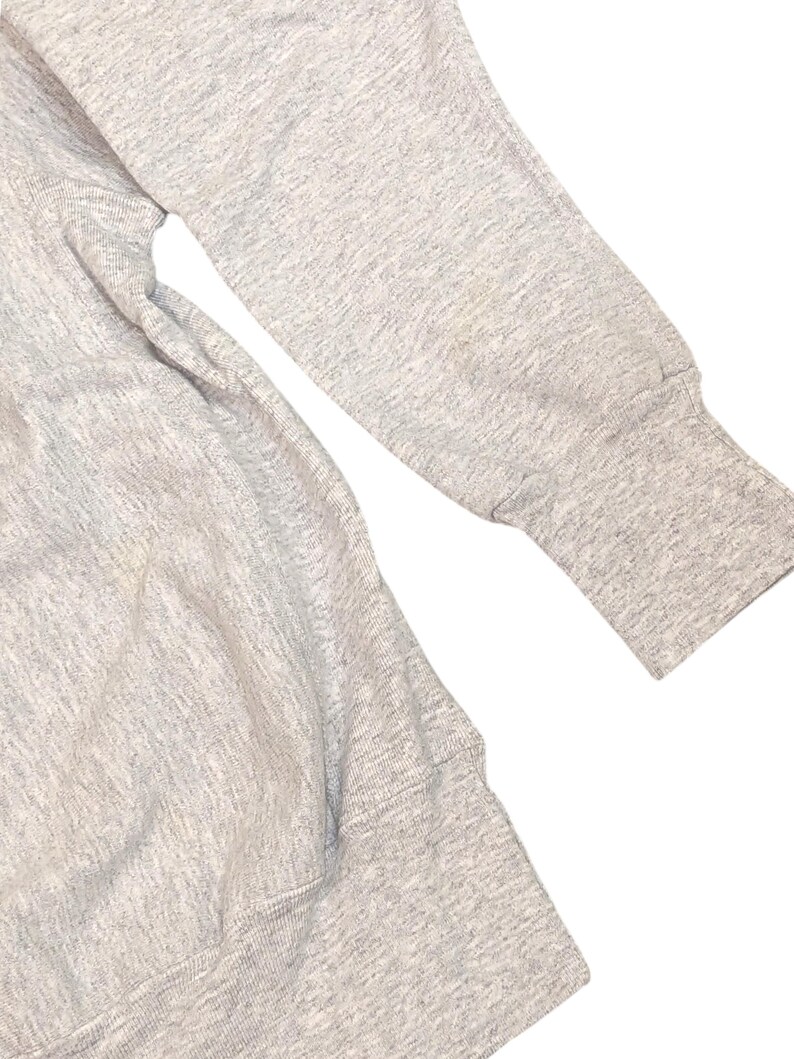 K&ouml;nnte beinhalten: Nahaufnahme eines hellgrauen Sweatshirt-&Auml;rmels und -K&ouml;rpers. Der &Auml;rmel hat ein geripptes B&uuml;ndchen und einen kleinen Fleck. Der Stoff scheint ein weiches, strukturiertes Material zu sein, wahrscheinlich Baumwolle oder eine Baumwollmischung. Das Sweatshirt ist ein Freizeitkleidungsst&uuml;ck.