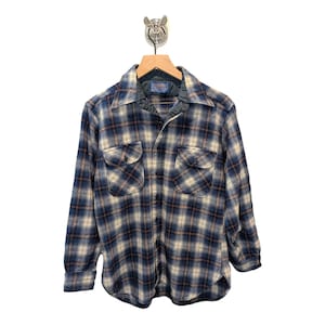 Pu&ograve; includere: Una camicia di flanella a quadri blu, beige e marrone con due tasche sul petto e una chiusura con bottoni. La camicia &egrave; appesa a una gruccia di legno con un gancio di metallo. La camicia ha un colletto e maniche lunghe.