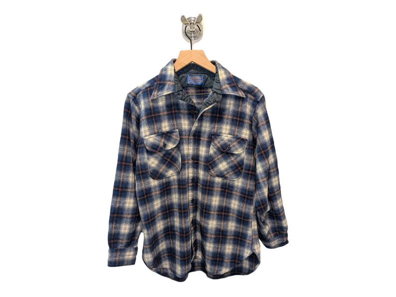 Pu&ograve; includere: Una camicia di flanella a quadri blu, beige e marrone con due tasche sul petto e una chiusura con bottoni. La camicia &egrave; appesa a una gruccia di legno con un gancio di metallo. La camicia ha un colletto e maniche lunghe.