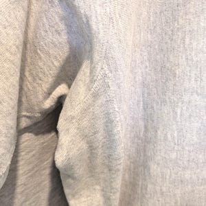 May include: Close-up of a light gray sweatshirt. The fabric has a textured appearance with visible stitching along the seams. The image highlights the shoulder and upper sleeve area, showcasing the garment's construction.