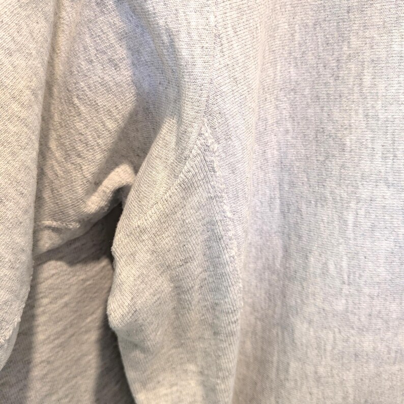 May include: Close-up of a light gray sweatshirt. The fabric has a textured appearance with visible stitching along the seams. The image highlights the shoulder and upper sleeve area, showcasing the garment's construction.