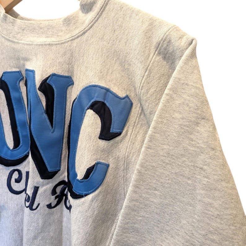 May include: A light gray sweatshirt with the letters "UNC" in light blue and black. The sweatshirt has a crew neck and a ribbed collar. The letters are appliqued onto the front of the sweatshirt.