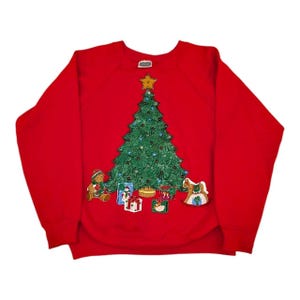 May include: A red Christmas sweatshirt featuring a green Christmas tree with a gold star on top. The tree is decorated with ornaments and lights. Presents and a teddy bear are at the base of the tree. A rocking horse is also present.