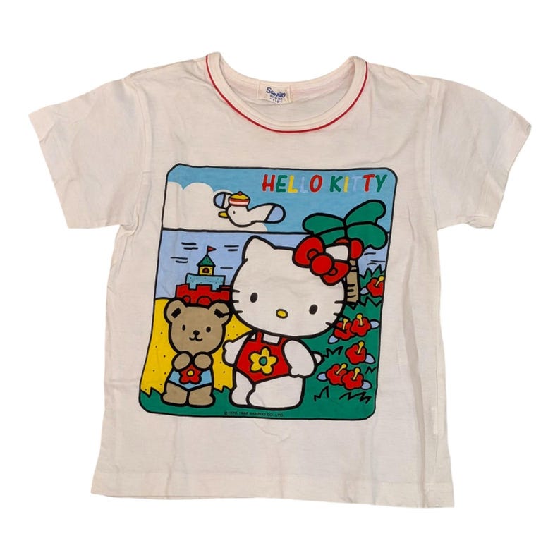 以下が含まれることがあります： 赤いトリムが襟ぐりに施された白いTシャツ。ハローキティのグラフィックが特徴です。グラフィックには、ハローキティ、テディベア、カラフルな文字で「HELLO KITTY」と書かれたビーチシーンが描かれています。