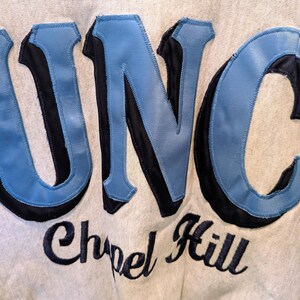 May include: Close-up of a light gray sweatshirt with the letters "UNC" in light blue and black, with "Chapel Hill" embroidered below in black script. The sweatshirt has a vintage, collegiate aesthetic.