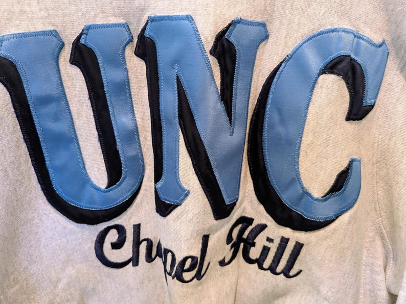 May include: Close-up of a light gray sweatshirt with the letters "UNC" in light blue and black, with "Chapel Hill" embroidered below in black script. The sweatshirt has a vintage, collegiate aesthetic.