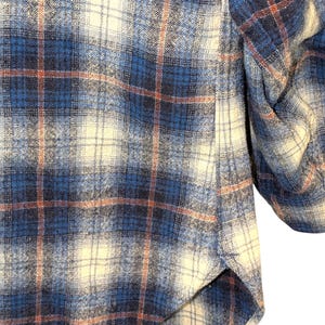 Pu&ograve; includere: Primo piano di una camicia di flanella a quadri blu, crema e ruggine. Il tessuto ha un aspetto morbido e strutturato. Il design della camicia presenta un classico motivo a quadri, con un polsino abbottonato visibile.