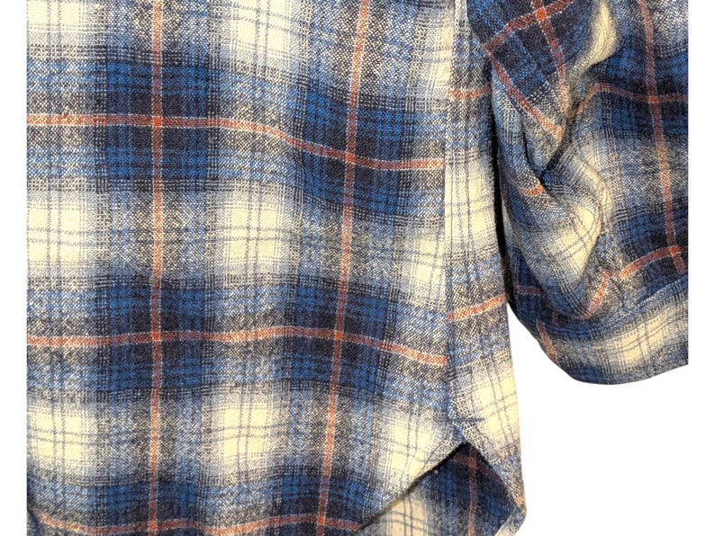 Pu&ograve; includere: Primo piano di una camicia di flanella a quadri blu, crema e ruggine. Il tessuto ha un aspetto morbido e strutturato. Il design della camicia presenta un classico motivo a quadri, con un polsino abbottonato visibile.