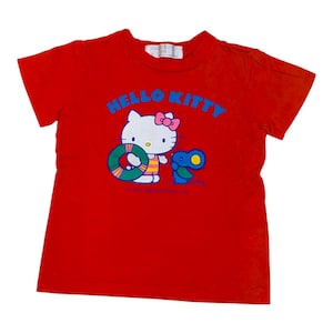 May include: Red t-shirt featuring a Hello Kitty graphic. The design includes Hello Kitty with a pink bow, a life preserver, and a blue creature. The words "HELLO KITTY" are in blue across the top.