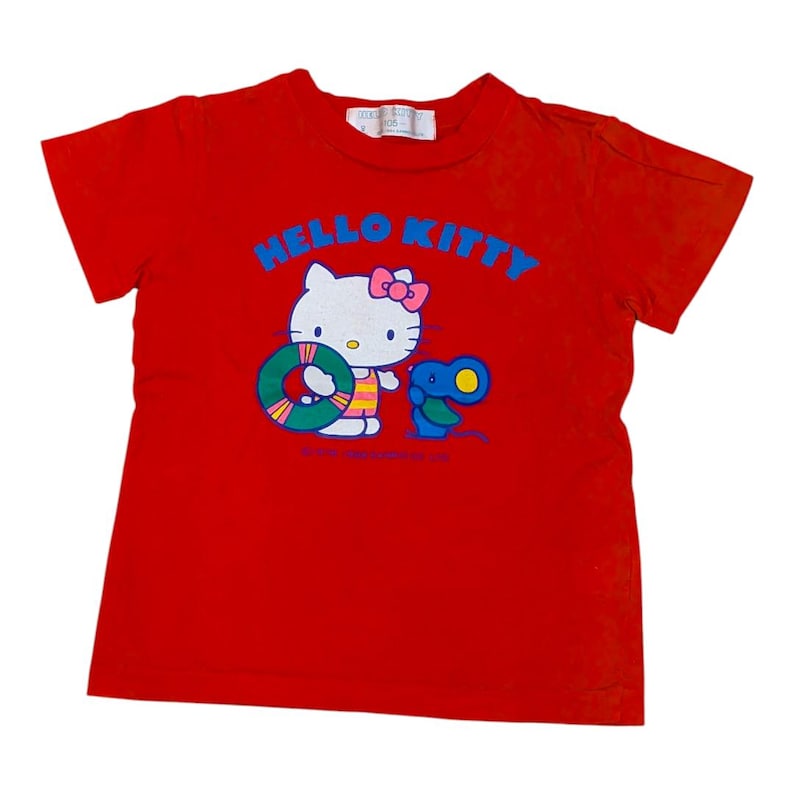 May include: Red t-shirt featuring a Hello Kitty graphic. The design includes Hello Kitty with a pink bow, a life preserver, and a blue creature. The words "HELLO KITTY" are in blue across the top.