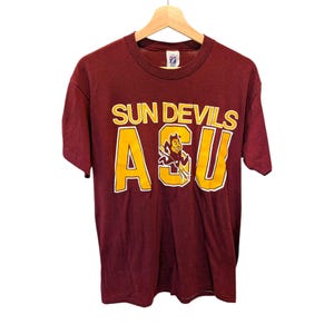 May include: A maroon short-sleeve t-shirt featuring the text SUN DEVILS in yellow above the large yellow letters ASU. The letter S in ASU contains a graphic of a cartoon devil character holding a pitchfork. The shirt is displayed on a hanger.