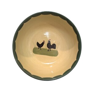May include: A round ceramic bowl with a cream-colored interior and a dark green scalloped rim. The center of the bowl features a painted illustration of two black chickens standing on a green patch of grass with thin blades of grass.