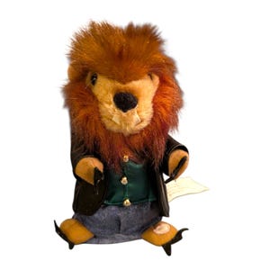 May include: A plush animal figure with a brown faux fur mane, tan face, and black nose. It wears a black jacket over a green vest with three silver buttons, and blue fabric trousers. The figure has small black claws and a white tag attached to its side.