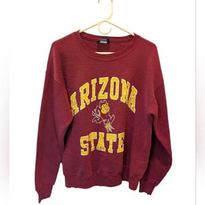 Puede incluir: Sudadera burdeos de cuello redondo con las palabras "ARIZONA STATE" en letras doradas. Una mascota de diablo dorada está centrada entre las palabras. La sudadera está colgada de una percha blanca.