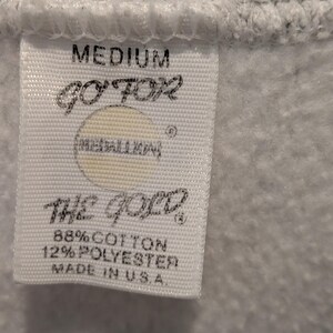 May include: Close-up of a white tag with black text, attached to a gray garment. The tag reads "MEDIUM", "GO FOR", "THE GOLD", and "88% COTTON 12% POLYESTER MADE IN U.S.A."