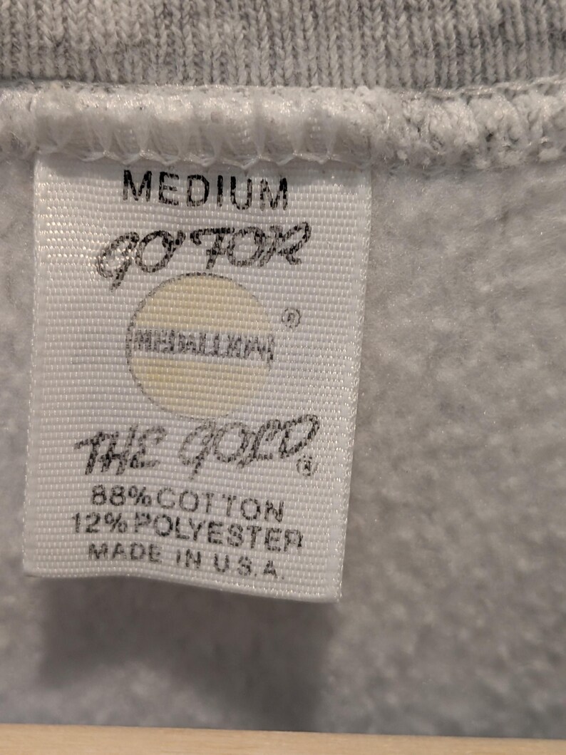 May include: Close-up of a white tag with black text, attached to a gray garment. The tag reads "MEDIUM", "GO FOR", "THE GOLD", and "88% COTTON 12% POLYESTER MADE IN U.S.A."