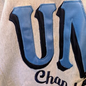 May include: Close-up of a light gray sweatshirt with the word "UNC" in large, light blue, and black stitched letters. Below the letters, the word "Chapel" is embroidered in a dark blue script.
