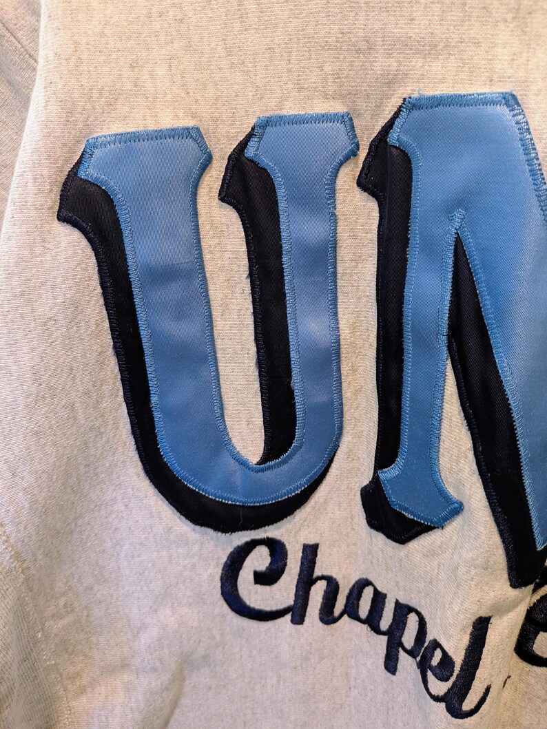 May include: Close-up of a light gray sweatshirt with the word "UNC" in large, light blue, and black stitched letters. Below the letters, the word "Chapel" is embroidered in a dark blue script.