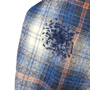 Pu&ograve; includere: Primo piano di un tessuto a quadri blu, bianco e arancione con un motivo ricamato blu scuro. Il tessuto ha un aspetto morbido e strutturato, che suggerisce una flanella o un materiale tessuto. Il ricamo aggiunge un elemento unico e artigianale.
