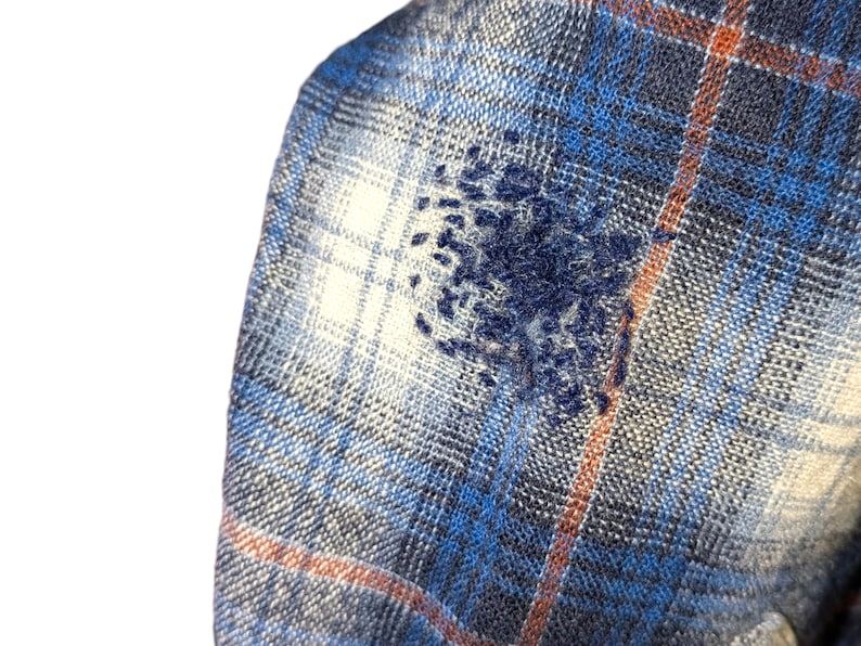 Pu&ograve; includere: Primo piano di un tessuto a quadri blu, bianco e arancione con un motivo ricamato blu scuro. Il tessuto ha un aspetto morbido e strutturato, che suggerisce una flanella o un materiale tessuto. Il ricamo aggiunge un elemento unico e artigianale.