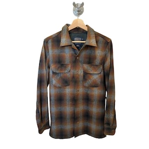 Puede incluir: Camisa abotonada a cuadros en tonos marrón, negro y gris con dos bolsillos en el pecho y mangas largas. La camisa está colgada de una percha de madera. Estilo clásico.