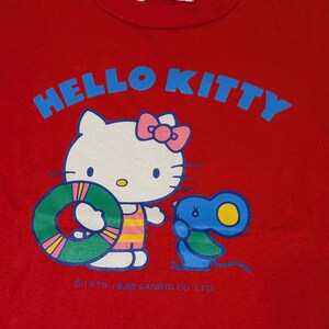 May include: Red Hello Kitty t-shirt featuring a graphic of Hello Kitty holding a green and yellow floatie, with a blue mouse. The words "HELLO KITTY" are in blue, above the graphic. The shirt has a white tag at the neckline.