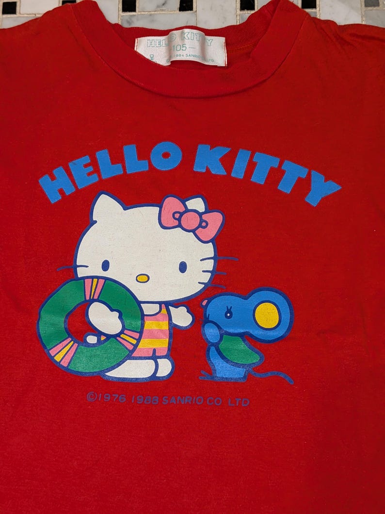 May include: Red Hello Kitty t-shirt featuring a graphic of Hello Kitty holding a green and yellow floatie, with a blue mouse. The words "HELLO KITTY" are in blue, above the graphic. The shirt has a white tag at the neckline.
