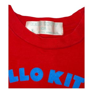 May include: A red t-shirt with the words "HELLO KITTY" in blue block letters. The shirt has a crew neck and a tag with the words "HELLO KITTY" and "105" printed on it.