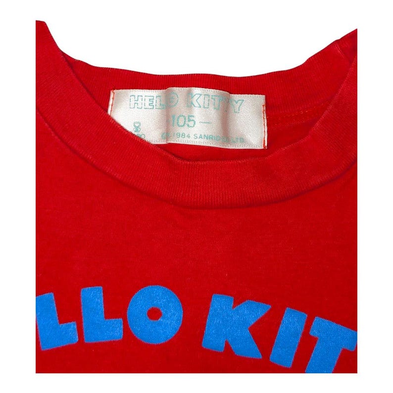 May include: A red t-shirt with the words "HELLO KITTY" in blue block letters. The shirt has a crew neck and a tag with the words "HELLO KITTY" and "105" printed on it.