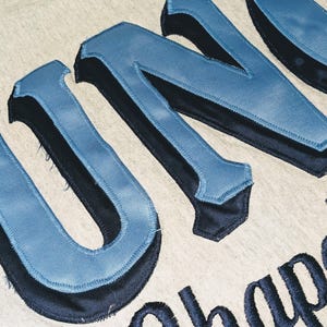 K&ouml;nnte beinhalten: Nahaufnahme eines hellgrauen Stoffes mit gro&szlig;en, blau und schwarz gestickten Buchstaben, die "UNO" und das Wort "Chap" buchstabieren. Die Buchstaben haben ein Satin-Finish und einen 3D-Effekt.