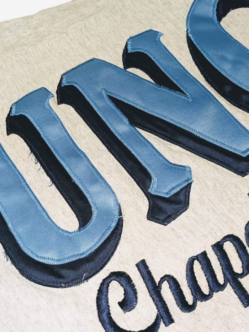 K&ouml;nnte beinhalten: Nahaufnahme eines hellgrauen Stoffes mit gro&szlig;en, blau und schwarz gestickten Buchstaben, die "UNO" und das Wort "Chap" buchstabieren. Die Buchstaben haben ein Satin-Finish und einen 3D-Effekt.
