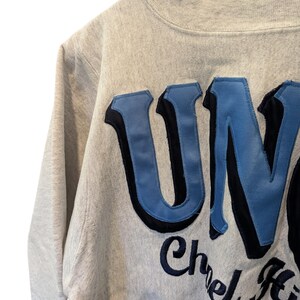 May include: Close-up of a light gray sweatshirt with the word "UN" in large, light blue letters outlined in black. The word "Chel" is visible in navy blue script below the letters.