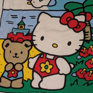 以下が含まれることがあります： トロピカルな環境で、ハローキティとテディベアのカラフルなグラフィックが描かれた白いTシャツ。デザインには「HELLO KITTY」の文字が入った青い空とビーチのシーンが含まれています。著作権情報も表示されています。