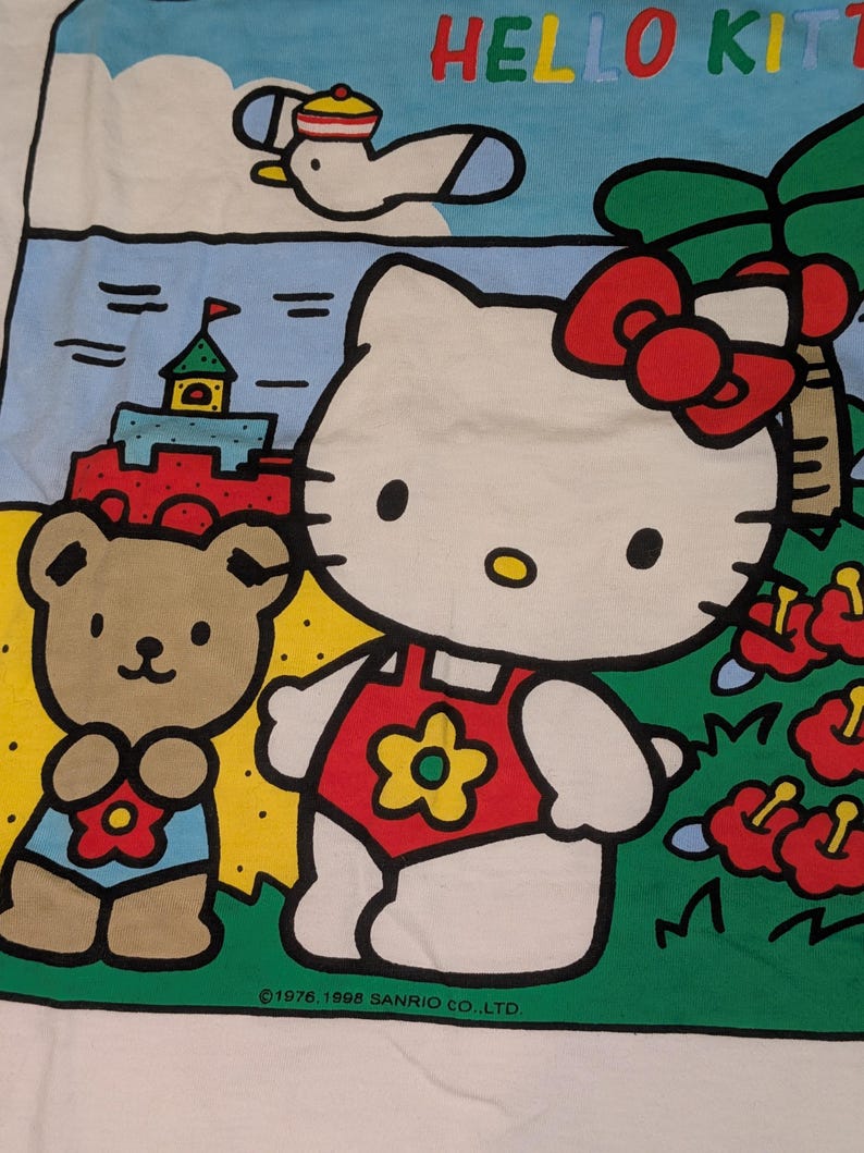 以下が含まれることがあります： トロピカルな環境で、ハローキティとテディベアのカラフルなグラフィックが描かれた白いTシャツ。デザインには「HELLO KITTY」の文字が入った青い空とビーチのシーンが含まれています。著作権情報も表示されています。