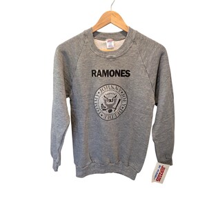 May include: A heather gray long-sleeve sweatshirt featuring the black text RAMONES above a circular graphic. The graphic depicts a seal with an eagle, a baseball bat, and the names Tommy, Johnny, Joey, and Dee Dee. A Jerzees by Russell tag is attached.