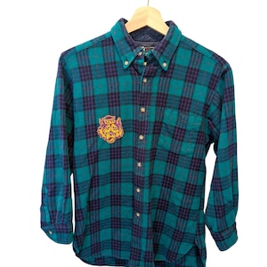 Puede incluir: Una camisa a cuadros verde y azul marino con un parche de tigre amarillo y naranja. La camisa de manga larga está colgada de una percha de madera. La camisa tiene cuello abotonado y un bolsillo en el pecho.