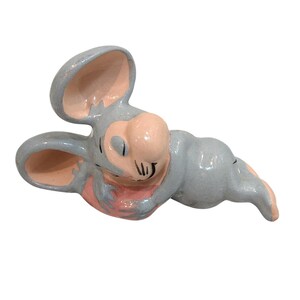 May include: A ceramic mouse figurine in a playful pose. The mouse is primarily light gray with pink accents on the ears, nose, and paws. The figurine has a glossy finish and is set against a white background.