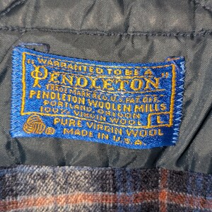 Pu&ograve; includere: Primo piano di un tessuto a fantasia scozzese blu e arancione con un'etichetta blu. L'etichetta recita "PENDLETON" e "PURE VIRGIN WOOL" con la taglia "L". Il tessuto &egrave; prodotto negli Stati Uniti.
