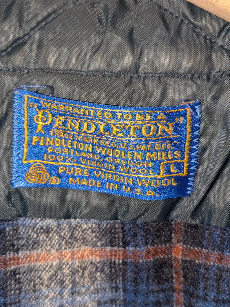 Pu&ograve; includere: Primo piano di un tessuto a fantasia scozzese blu e arancione con un'etichetta blu. L'etichetta recita "PENDLETON" e "PURE VIRGIN WOOL" con la taglia "L". Il tessuto &egrave; prodotto negli Stati Uniti.
