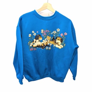 May include: A blue sweatshirt with a white collar and a graphic of two puppies and a kitten playing among flowers. The puppies are brown and black, and the kitten is orange and brown. The flowers are pink, white, and yellow.