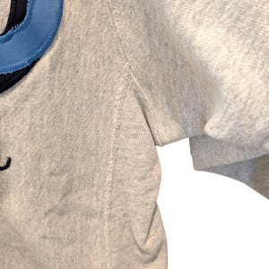 May include: Close-up of a light gray sweatshirt with a blue and navy design on the upper left. The sleeve is visible on the right side. The fabric has a textured appearance.
