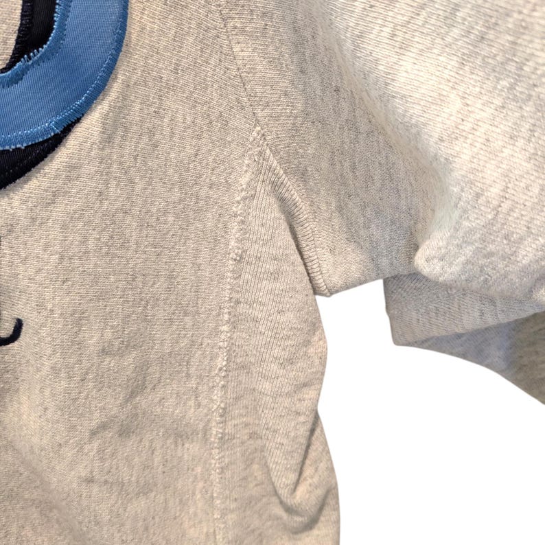 May include: Close-up of a light gray sweatshirt with a blue and navy design on the upper left. The sleeve is visible on the right side. The fabric has a textured appearance.