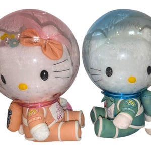 May include: Two plush Hello Kitty dolls wearing astronaut suits. One doll has a pink helmet and orange suit, the other has a blue helmet and teal suit. Both have clear bubble helmets.