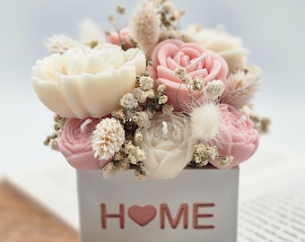 Könnte beinhalten: Ein floraler Kerzenarrangement in einem quadratischen Behälter mit dem ausgeschnittenen Wort "HOME". Das Arrangement zeigt weiße und rosa rosenförmige Kerzen, Trockenblumen und einen kleinen Kerzendocht in jeder Blume.