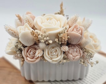 Könnte beinhalten: Ein Blumenarrangement aus cremefarbenen und rosafarbenen rosenförmigen Kerzen und Trockenblumen in einem weißen, geriffelten rechteckigen Behälter. Die Kerzen sind so angeordnet, dass sie einem Bouquet ähneln. Der Behälter steht auf einem Buch.