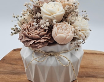 Könnte beinhalten: Ein dekoratives Blumenarrangement aus blütenförmigen Kerzen in einer weißen geometrischen Vase. Die Kerzen sind in verschiedenen Creme-, Pfirsich- und Mauve-Tönen gehalten, mit Schleierkraut. Eine goldfarbene Schnur-Schleife verziert die Vase.