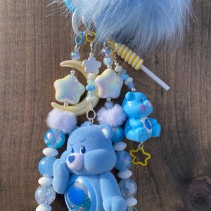 May include: A light blue Care Bear-themed keyring with a fluffy blue pom-pom, star and moon charms, and a lollipop. The keyring features a large Care Bear figure and a smaller Care Bear figure, both in blue.