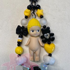 Puede incluir: Un llavero hecho a mano con una pequeña muñeca con un sombrero amarillo en forma de maíz, rodeada de cuentas negras, amarillas y blancas, un lazo negro, un dije de oso negro y un dije de corazón amarillo. El llavero tiene un cierre negro.
